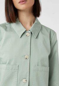Jasnozielona kurtka zapinana na guziki, wykonana z gładkiego materiału, z kołnierzykiem, dwoma kieszeniami na piersi i białymi guzikami.