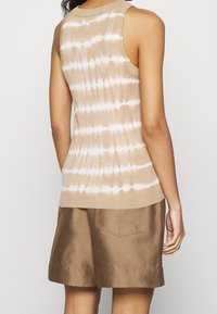 Béžový žebrovaný tílko s bílým batikovaným vzorem, s přiléhavým střihem a širokými výstřihy na paže, doplněné hnědými šortkami s kapsami.