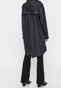 Manteau noir imperméable avec capuche, présentant un design épuré, une coupe longue et une texture lisse. Un pantalon noir évasé complète le look.