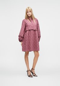 Φόρεμα με βολάν στο τελείωμα από υφή μοβ ύφασμα, με μακριά μανίκια και γιακά με κουμπιά. Συνδυασμένο με μαύρα ψηλοτάκουνα σανδάλια με λουράκια.