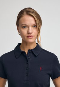 Navyblaue Poloshirt aus strukturiertem Stoff, mit einem klassischen Kragen, sechs Knöpfen und einem kleinen roten Logo auf der linken Brust.