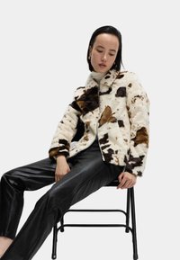 Umělá kožešinová bunda s kravským vzorem v bílé, hnědé a černé barvě. Zapínání na zip, měkká textura a límeček. Nositá přes černé kožené kalhoty.