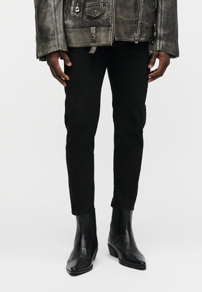 Personne à la peau foncée portant un pantalon noir ajusté, des bottines en cuir noir et une veste en cuir grise usée, se tenant devant un fond uni.