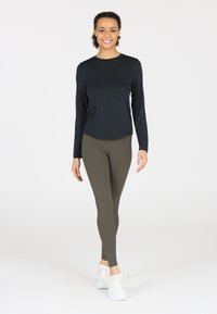 Zwarte lange mouwen top gecombineerd met olijfgroene leggings. Beide kledingstukken hebben een gladde textuur. Model draagt witte sneakers.