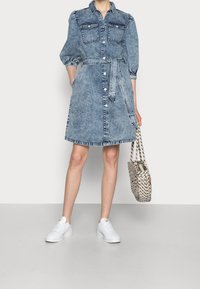 Nainen, jolla on vyöllinen, nappikiinnitteinen farkkukleid, puffihihat, valkoiset sneakerit ja jossa on kuvioitu olkalaukku yhdessä kädessä.