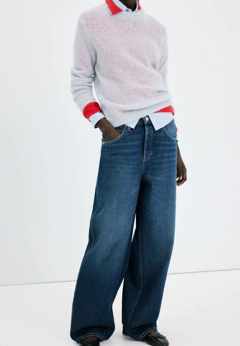Personne portant un jean en denim bleu surdimensionné, un pull en maille blanc, une chemise rouge à col en dessous, debout avec une main dans la poche.