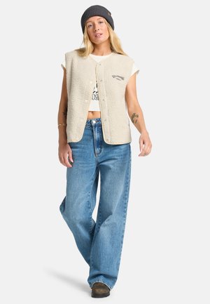 Beige fleece vest with short sleeves, snap button closure, and a logo detail. Paired with loose blue jeans and a gray beanie. Brown footwear.