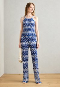 Combinaison à motifs zigzag bleus et blancs avec des bretelles fines et un buste ajusté, s'évase aux jambes, accompagnée d'un petit sac léger.