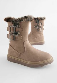 Botas de tobillo marrones hechas de un material similar al ante con un puño de pelo sintético. Presenta dos hebillas plateadas y una suela de goma texturizada.