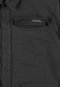 Czarna koszula z tkaniny z wyostrzonym kołnierzykiem, pojedynczą kieszenią na piersi i subtelnym logo na kieszeni. Teksturowany materiał o gładkim wykończeniu.