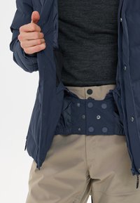 Marineblaue isolierte Jacke mit Reißverschluss, elastischem Saum und Druckknopfverschlüssen, getragen über einem dunkelgrauen Hemd und beigen Hosen mit glatter Oberfläche.