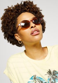 Óculos de sol redondos castanhos com uma armação em marrom, combinados com uma t-shirt amarelo claro com uma gráfica de palmeira. Cabelo texturizado adiciona volume.