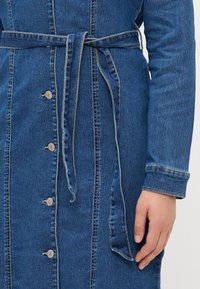 Σαλοπέτα από τζιν με ζώνη στη μέση, κουμπάκια εμπρός και μακριά μανίκια. Το ύφασμα είναι σκούρο μπλε με διακριτή υφή και αντίθεση ραφής.