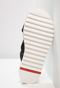 Sneaker nera con tomaia in pelle testurizzata, suola in gomma bianca con scanalature orizzontali e un accento rosso vicino al tallone.