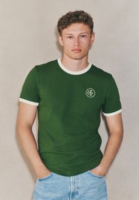 T-shirt di cotone verde con collo a giro e bordi delle maniche a costine bianchi. Presenta un piccolo logo sul petto a sinistra. Abbinata a pantaloncini corti in denim azzurro chiaro.