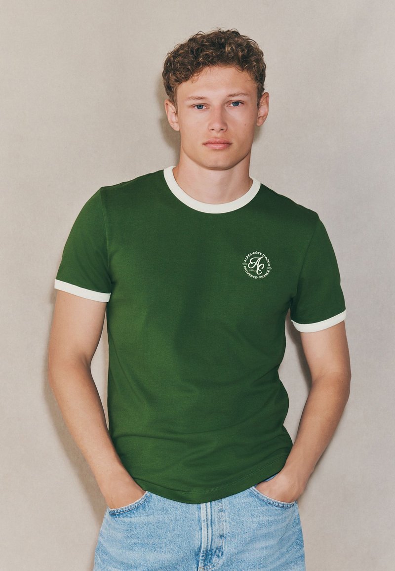 T-shirt di cotone verde con collo a giro e bordi delle maniche a costine bianchi. Presenta un piccolo logo sul petto a sinistra. Abbinata a pantaloncini corti in denim azzurro chiaro.