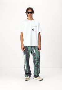 T-shirt en coton blanc avec une poche poitrine et un logo, associé à un pantalon à motifs verts, gris et blancs. Modèle portant des lunettes de soleil et des baskets.