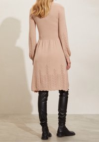 Långärmad, ljusrosa stickad klänning med en texturerad nederkant och geometriska mönster längst ner, kombinerad med svarta stövlar över knäna.