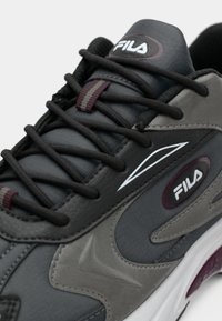Szare i czarne sneakersy z teksturowaną syntetyczną cholewką, grubymi sznurówkami i wyraźnym logo, z białą podeszwą środkową i bordowymi akcentami.