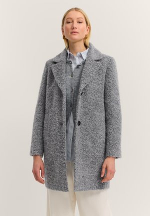 Frau trägt einen strukturierten grauen Wollmantel über einem grauen Knopfcardigan und einem blauen gestreiften Hemd, kombiniert mit weißen Hosen.