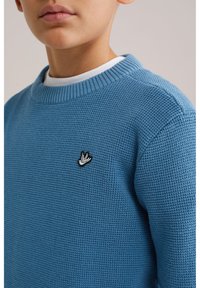 Pull bleu en tricot avec un col rond, présentant un motif texturé et un petit logo d'oiseau noir et blanc sur la poitrine.