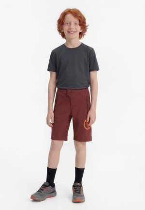 Nasmejan rdečelasi deček stoji, oblečen v temno sivo majico, bordo kratke hlače z oranžnim logotipom, črne nogavice in sive-oranžne superge na belem ozadju.