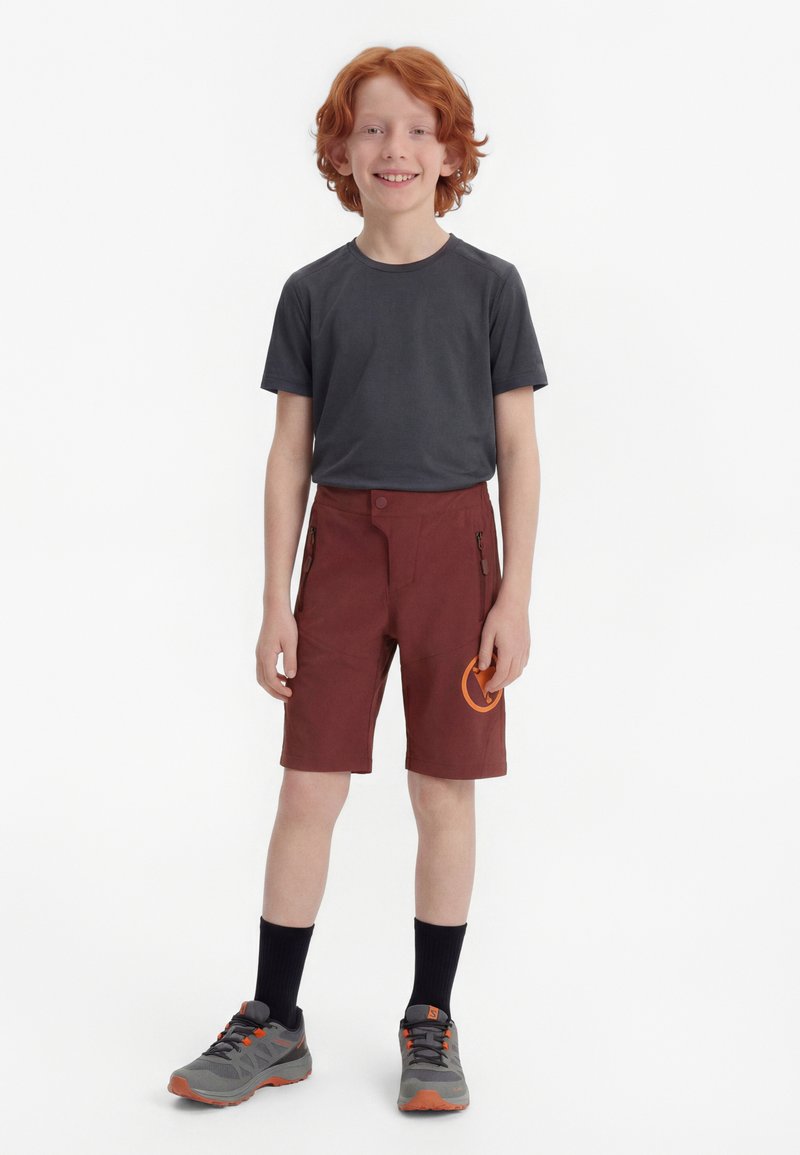 Χαμογελαστός αγόρι με κόκκινα μαλλιά που στέκεται, φοράει σκούρο γκρι μπλουζάκι, μπορντό σορτς με πορτοκαλί λογότυπο, μαύρες κάλτσες και γκρι-πορτοκαλί αθλητικά παπούτσια σε λευκό φόντο.