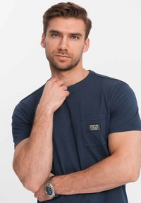Námořnická modrá tričko z měkké tkaniny, s kapsou na levé části hrudi a nášivkou loga. Krátké rukávy a klasický střih.