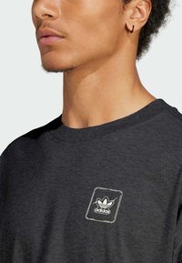 Černé bavlněné tričko s kulatým výstřihem, s bílou nášivkou s logem Adidas na levé straně hrudi a detailem z texturované látky.