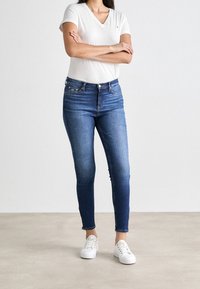 Jeans ajustados en denim oscuro con cintura alta, que presentan un diseño clásico de cinco bolsillos y dobladillo deshilachado. Combinados con una camiseta blanca de cuello en V y zapatillas blancas.
