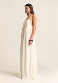 Robe maxi sans manches de couleur crème, avec une silhouette fluide, un tissu léger, des fentes latérales et un design minimaliste.