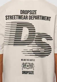 Cream-colored t-shirt featuring bold black text and graphic designs including stripes and a logo with lions, indicating a streetwear theme.