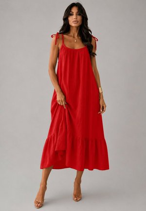 Femme debout portant une robe rouge ample sans manches avec des bretelles à nouer et des chaussures à talons transparents sur un fond gris uni.