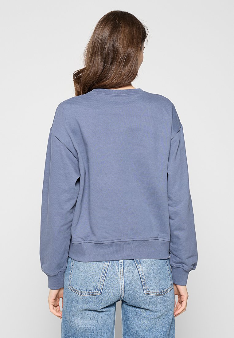 Felpa con girocollo aderente realizzata in morbido tessuto testurizzato di colore azzurro chiaro, con spalle scese e polsini a costine. La vista posteriore rivela un design semplice.