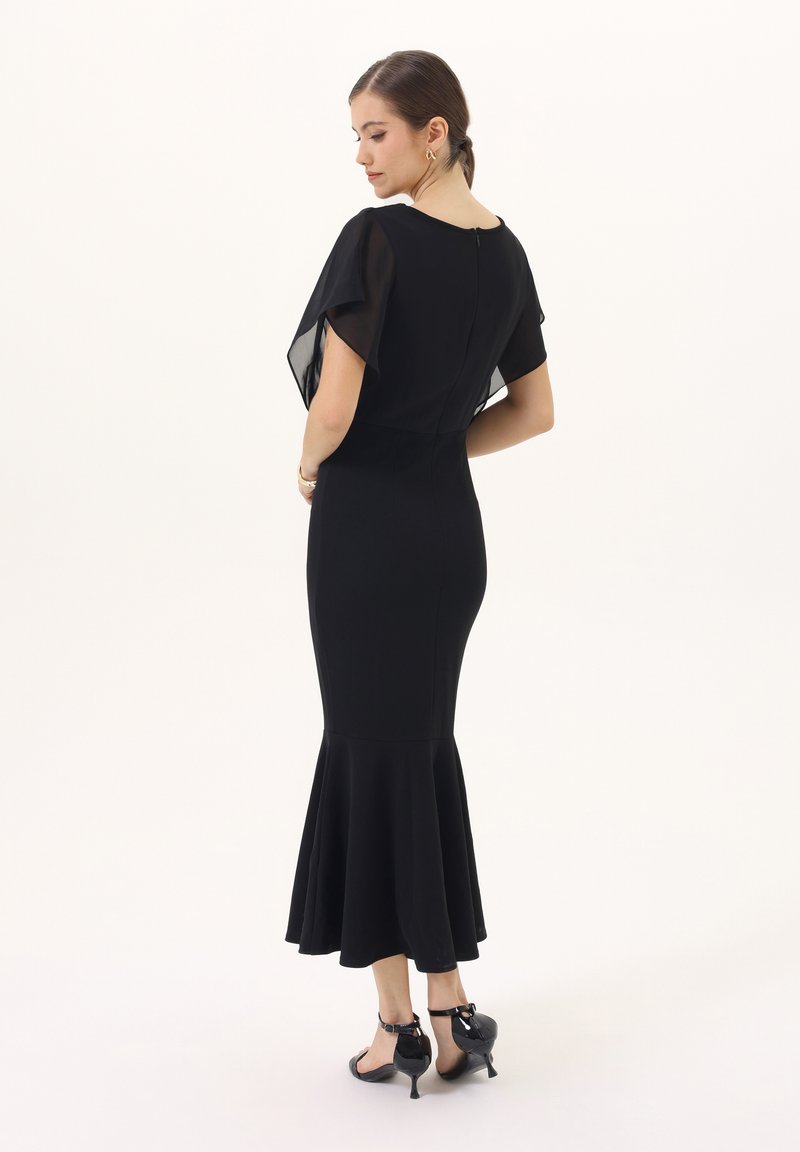 Czarna sukienka syrenka z przezroczystymi bufkami, dopasowanym gorsetem i rozkloszowanym dołem. Gładki materiał z zapięciem na suwak z tyłu. Czarne szpilki.