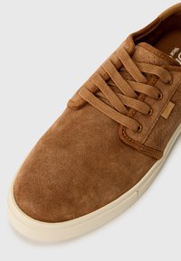 Zapatillas de ante marrón con una textura suave, que presentan cordones planos, perforaciones laterales y una suela de goma beis. Etiqueta con logo en el lateral.
