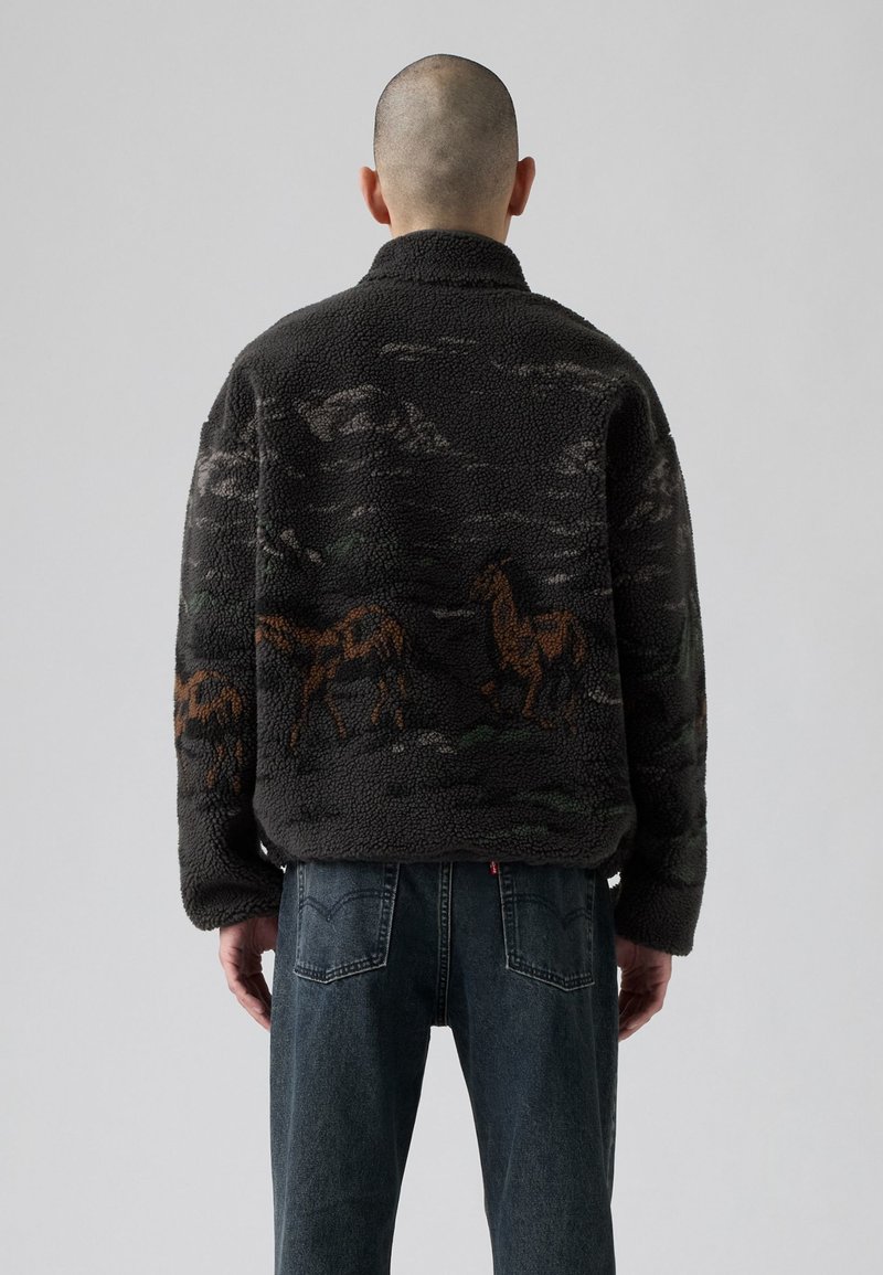 Mann mit rasiertem Kopf, der eine schwarze Fleecejacke mit braunen Pferdemotiven auf dem Rücken und dunkelblauen Jeans trägt, steht vor einem schlichten Hintergrund.