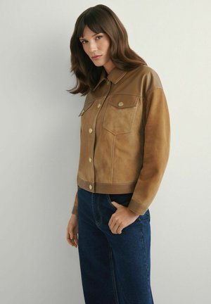 Kobieta o brązowych włosach, ubrana w beżową zamszową kurtkę z złotymi guzikami i niebieskie dżinsy, stojąca na jasnym, jednolitym tle.