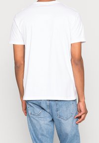 Bílý krátký rukáv T-shirt vyrobený z bavlny. Jednoduchý design s kulatým výstřihem, volnější střih. Nosený s světle modrými džínami, které mají zadní kapsy.
