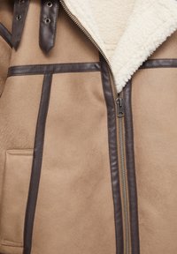Giacca di shearling color sabbia con dettagli in pelle marrone, dotata di chiusura con zip, bottoni a scatto e un interno morbido e testurizzato.