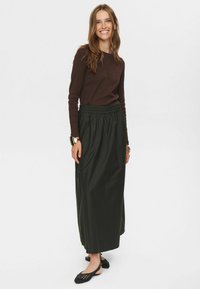 Jupe longue vert foncé avec une taille élastique, associée à un haut ajusté marron à manches longues. La femme porte des chaussures noires avec des détails.
