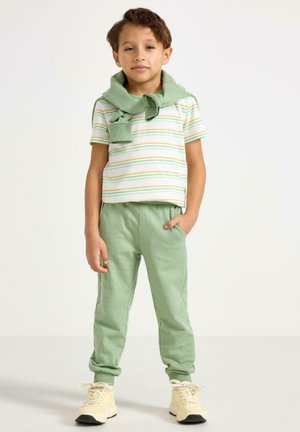 Vihreät collegehousut ja lyhythihainen raidallinen paita, jossa on keltaisia ja vihreitä viivoja; vaaleanvihreä neule solmittuna kaulan ympärille; valkoiset lenkkarit.