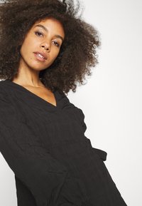 Vestido preto com decote em V, detalhes franzidos e mangas longas. Feito de tecido texturizado com um padrão subtil. Inclui um cinto de laço.