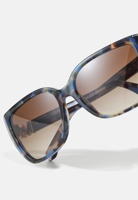 Sončna očala Michael Kors z velikim, prevelikim oblikovanjem mačjih oči. Okvir ima pisan barvit vzorec želve in gradientne rjave leče.