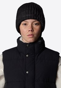 Czarna dziana czapka beanie z prążkowanym wzorem i szczegółowym logo, noszona z ciemną kamizelką o wysokim kołnierzu i srebrnymi zatrzaskami, na jasnej polarowej bluzie.