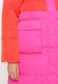 Giacca imbottita con sezioni arancioni e rosa, dotata di una grande tasca frontale e di una superficie liscia e lucida. Design leggero e isolante.