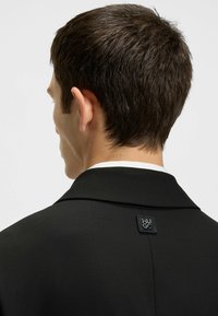 Black blazer with a smooth texture, featuring a pronounced collar and a rectangular logo patch on the back. Simple and tailored design.