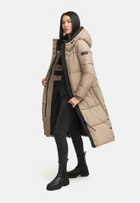 Beige lange gewatteerde jas met een capuchon, zwarte voering en drukknopen. Gekozen boven een zwarte top en strakke jeans, gecombineerd met zwarte laarzen.