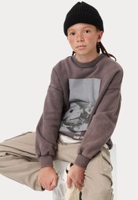 Felpa grigia con una grande stampa grafica, colletto rotondo e polsini a coste indossata sopra una camicia bianca, abbinata a pantaloni cargo beige e sneakers.