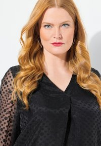 Top nero con pois texture, scollatura a V e maniche lunghe trasparenti. Il modello ha lunghi capelli mossi e pelle chiara. Labbra audaci completano il look.